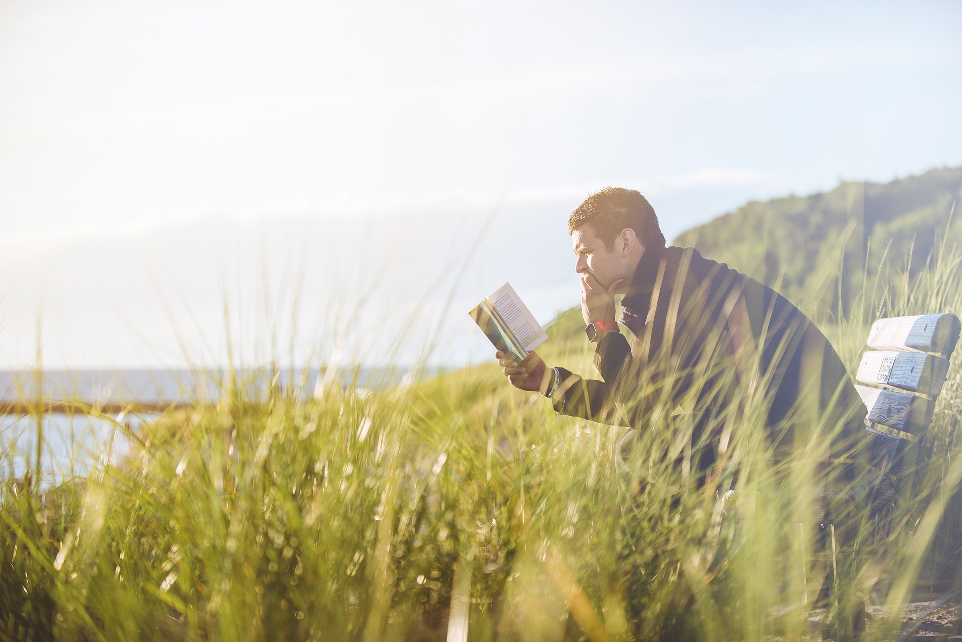 Foto: Mann liest ein Buch auf einer Bank in den Dünen, im Hintergrund das Meer